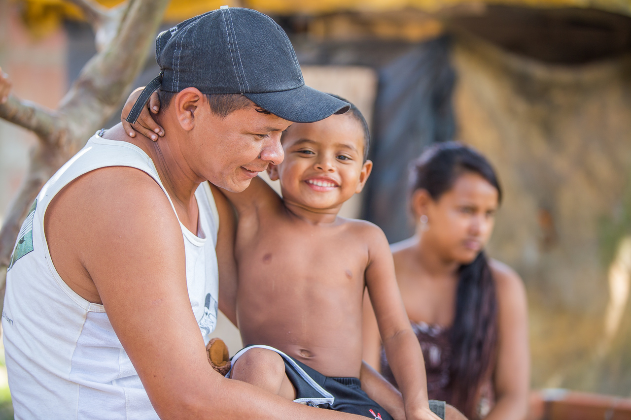 Na foto, um menino indígena está sentado no colo de seu pai e sorrindo. Seu pai, de blusa branca e boné, também sorri. Ao fundo, de forma desfocada, é possível ver uma mulher e residências.