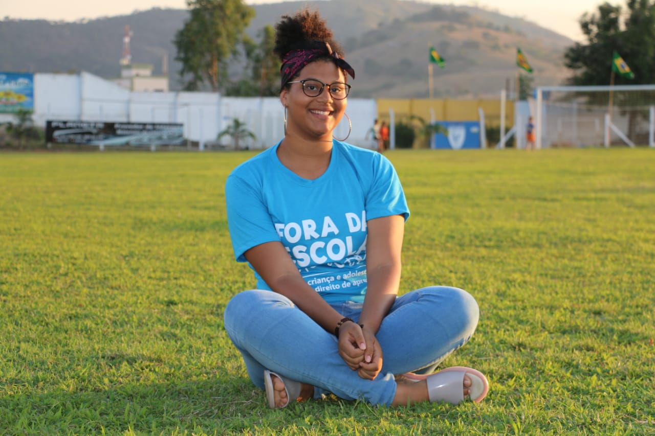 Adolescente sentada num gramado verde sorri para câmera. Ela veste calça jeans azul e uma blusa azul. Óculos de grau redondos, brincos de argola e uma faixa roxa e preta no cabelo 