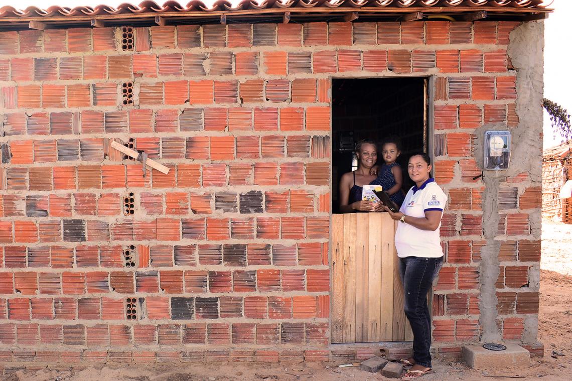 Uma agente, de blusa branca, está na porta de uma casa de tijolos. Ao seu lado está uma mulher negra jovem segurando seu filho, bebê, e sorrindo para a câmera