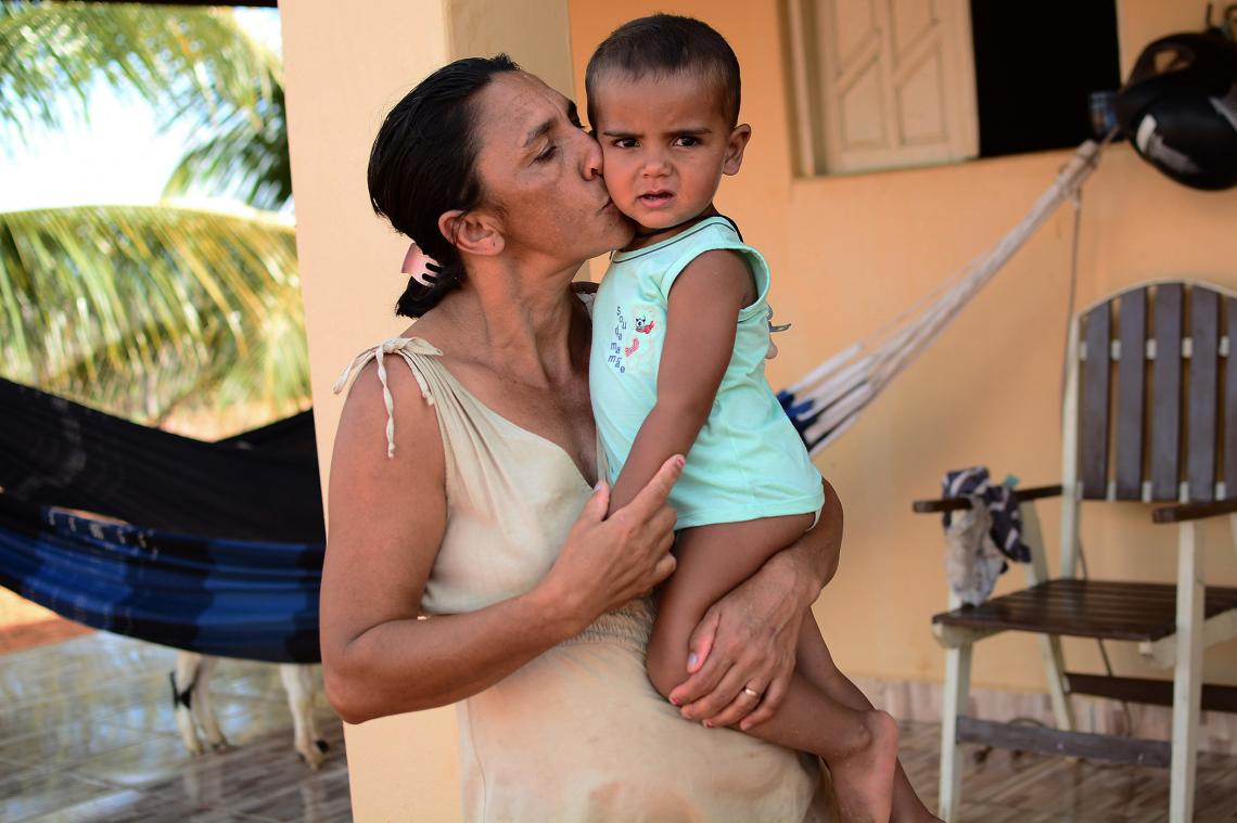 Rosivânia, uma mulher negra, segura seu filho bebê e o beija na bochecha. O menino olha em direção a câmera.