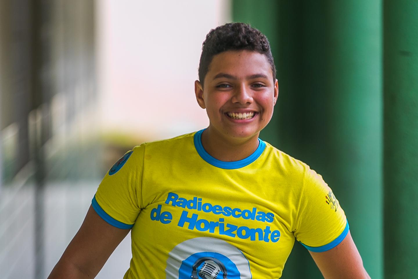 Adolescente sorrindo olha para a câmera. Veste camiseta amarela fosforescente com detalhes em azul na manga. A estampa diz: "Radioescolas de Horizonte"  