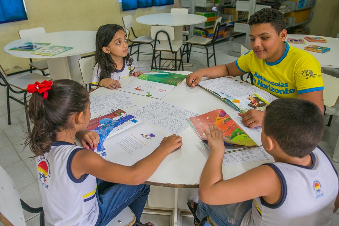 Acássio vestindo uma camiseta amarela fosforescente está sentado numa mesa redonda. Ao redor da mesa, três crianças menores de 12 anos vestem uniforme escolar e seguram livros nas mãos. A mesa está repleta de livros. Ao fundo, estantes com livros indicam que estão numa biblioteca escolar