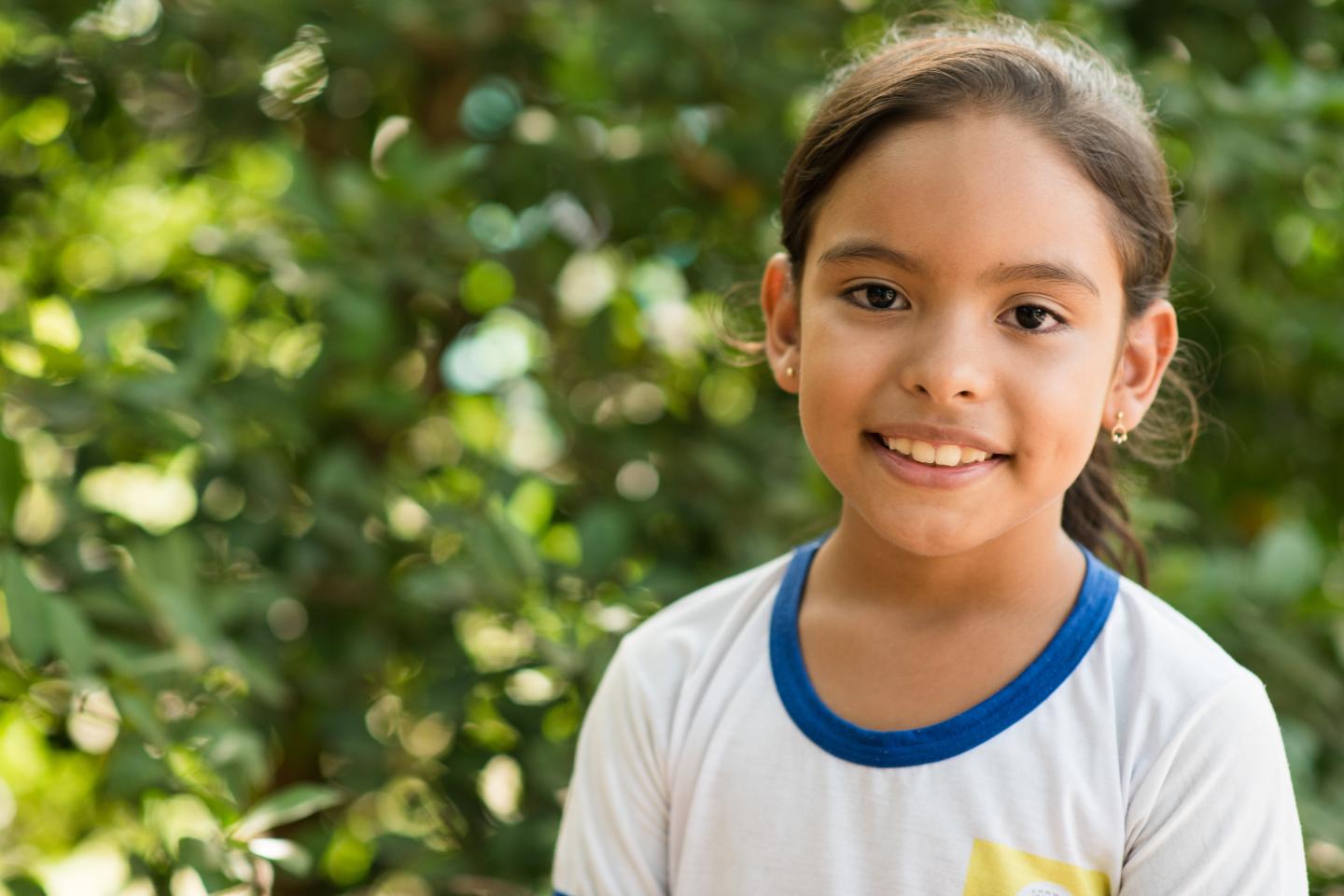 Criança com uniforme escolar branco de gola azul olha para a câmera e sorri. Ao fundo, temos uma vegetação verde