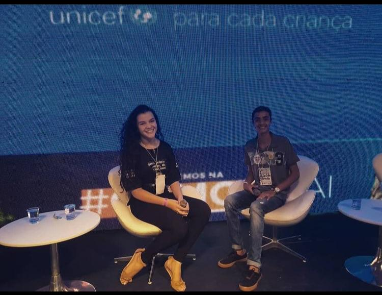 Adolescente vestindo blusa e calça preta sorri. Ela segura um microfone nas mãos e está sentada numa cadeira em cima de um palco. ao fundo, um painel do UNICEF. Ao lado dela, outro jovem sentado numa cadeira sorri