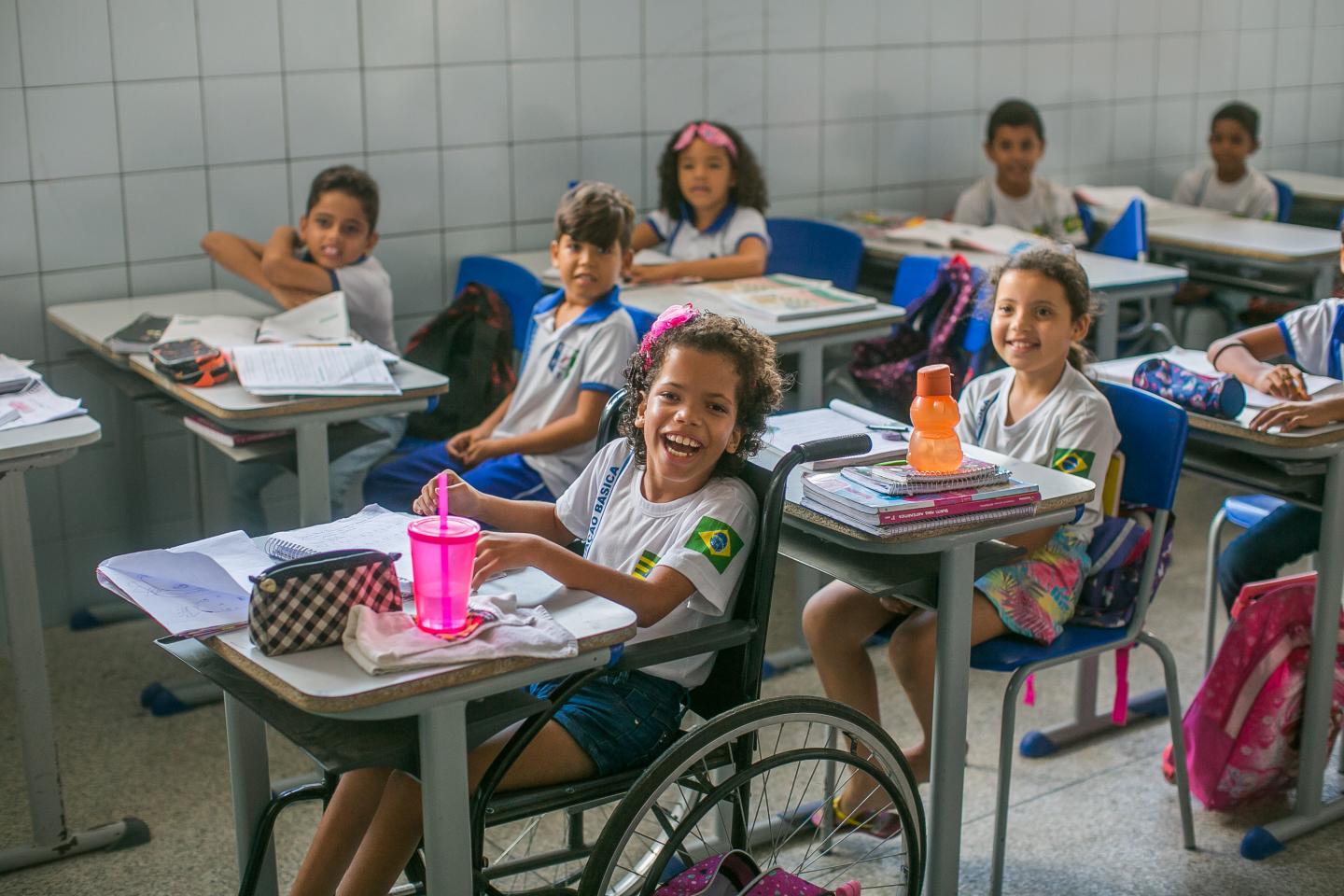 Vestindo uniforme escolar, menina sorri para câmera. Ela está numa cadeira de rodas encaixada na carteira escolar. Sobre a mesa, um estojo quadriculado preto e branco, uma garrafinha de água rosa e um caderno. Ao fundo da menina, outras crianças estão sentadas em suas carteiras vestindo uniforme escolar e olhando para frente