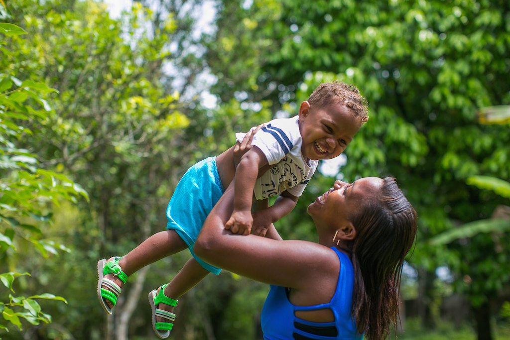 Mulher quilombola veste blusa azul e sorri para o filho pequeno. Ela o ergue. O menininho sorri, suspenso no ar. Ele veste uma blusa branca com listras azuis escuras e um desenho amarelo no peito, bermuda azul claro e sandalhas verdes fosforescentes. Ao fundo, uma vegetação verde