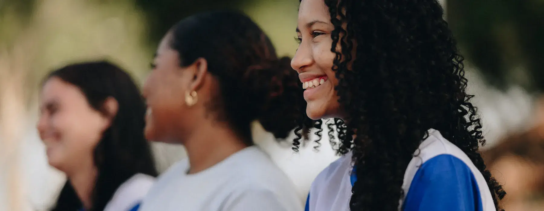 três meninas adolescentes sorriem, elas estão de perfil.