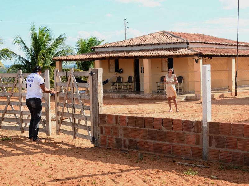 Uma agente comunitária, de blusa branca, está em pé no portão de uma casa no sertão de Pernambuco
